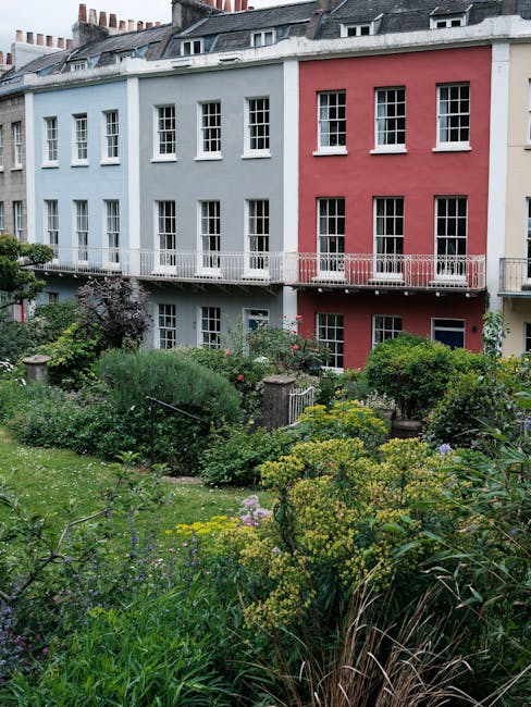 A charming urban garden in front of a row of multi-story residential buildings painted in light blue and pink. The garden features lush, varied plantings including flowering shrubs, ground cover plants, and ornamental grasses creating a vibrant, natural landscape. The soil appears moist and dark, indicating recent watering or rain. Pathways and garden beds are outlined with small stone or brick borders, with some areas slightly elevated. A metal railing runs along the edge of a balcony on the upper floor, overlooking the garden below. The scene is illuminated by soft natural light typical of a cloudy day, emphasizing the diverse textures of leaves and flowers. The overall impression is of a well-maintained, inviting outdoor space designed for aesthetic appeal and outdoor enjoyment, with visible gardening tools and careful pruning suggesting active garden maintenance by professionals such as Gardeners Putney.