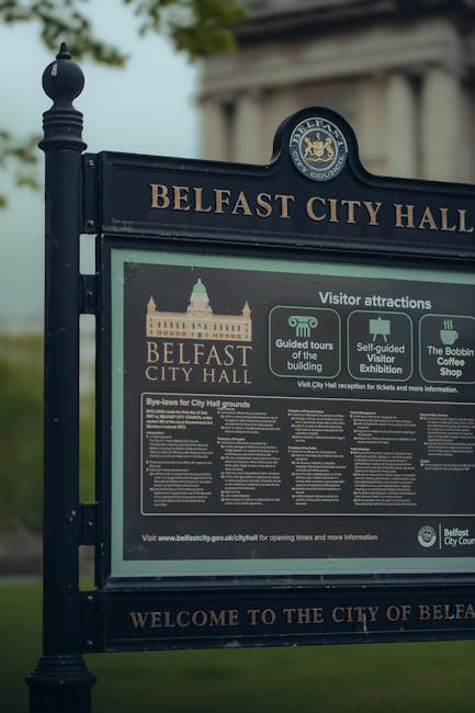 A close-up view of a large black informational sign outside Belfast City Hall, featuring gold lettering and graphics. The sign displays the words 'Belfast City Hall' along with the Belfast City Hall emblem at the top, and provides details about visitor attractions, guided tours of the building, self-guided visitor exhibitions, and information about the Bobbin Coffee Shop. Behind the sign, there are lush green trees with dense foliage, and a portion of a classical-style white columned building is visible in the background. The scene is lit by soft, natural daylight, highlighting the textures of the sign’s metal frame and the vibrant greenery surrounding the area. The overall atmosphere suggests a well-maintained outdoor space suitable for garden maintenance and landscaping work, typical of urban park settings overseen by city authorities like Belfast City Council.